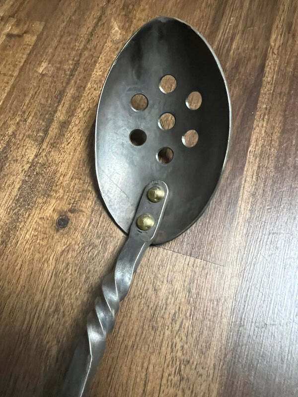 Hand forged strainer spoon with brass rivets on wooden surface.