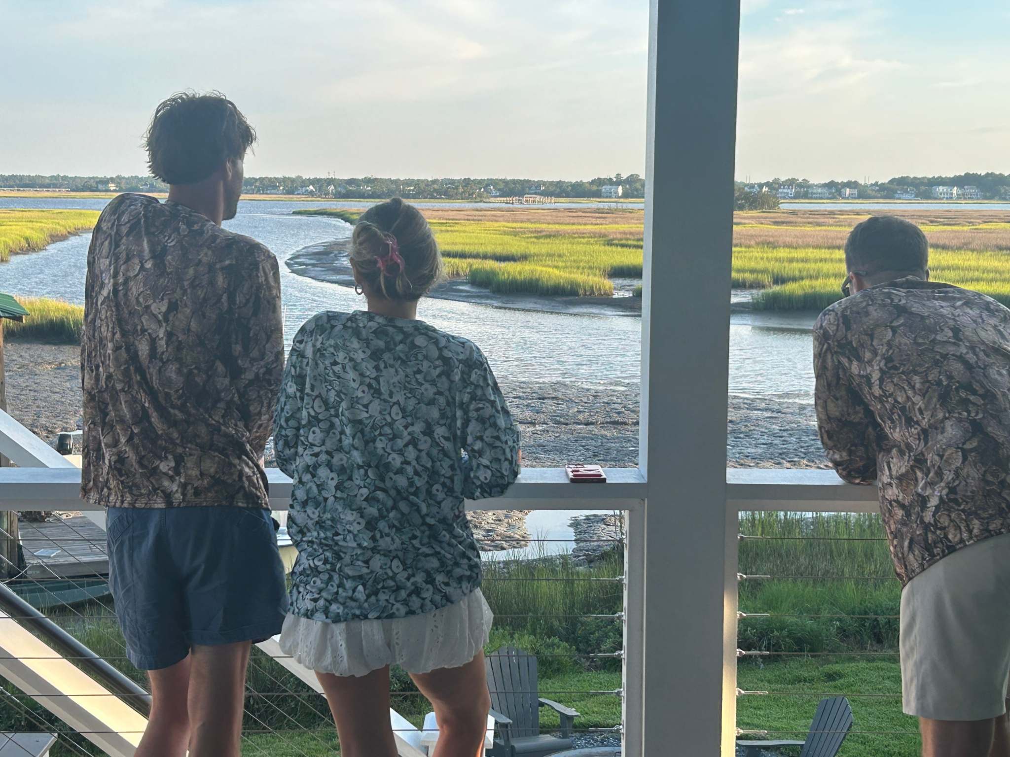Three people wearing Oystaflage Blue Point Long Sleeve Performance Shirts overlooking a coastal marsh at sunset