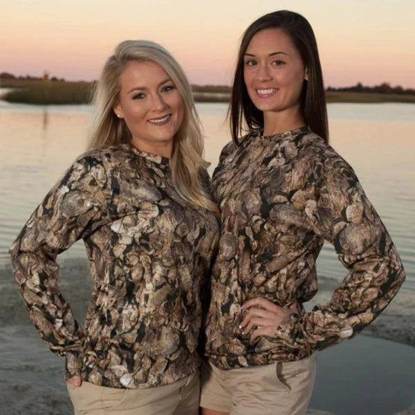 Two women wearing Oystaflage Long Sleeve camo performance shirts standing by the water at sunset